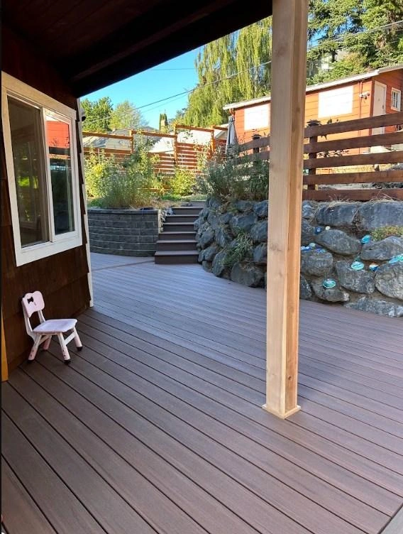 View From A Dark Composite Porch Over A Detailed Backyard Featuring Steps, A Stone Wall, And Garden Landscaping After A Rotted Wood Deck Replacement.
