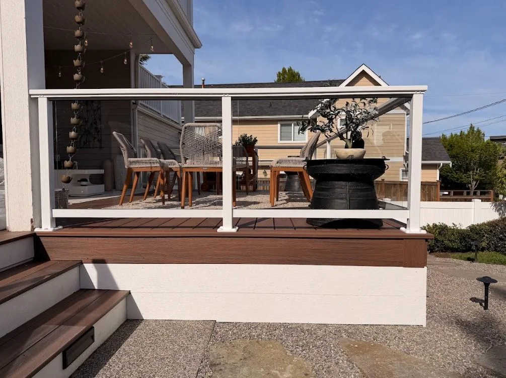 A Raised Patio Area Showcasing A New Ground-Level Azek Deck Replacement In Edmonds With White Glass Railings And Seating.